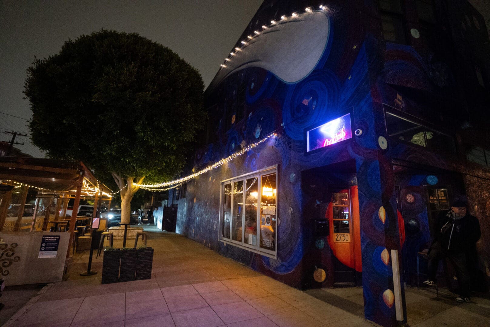 Nighttime street view with illuminated building and outdoor seating.